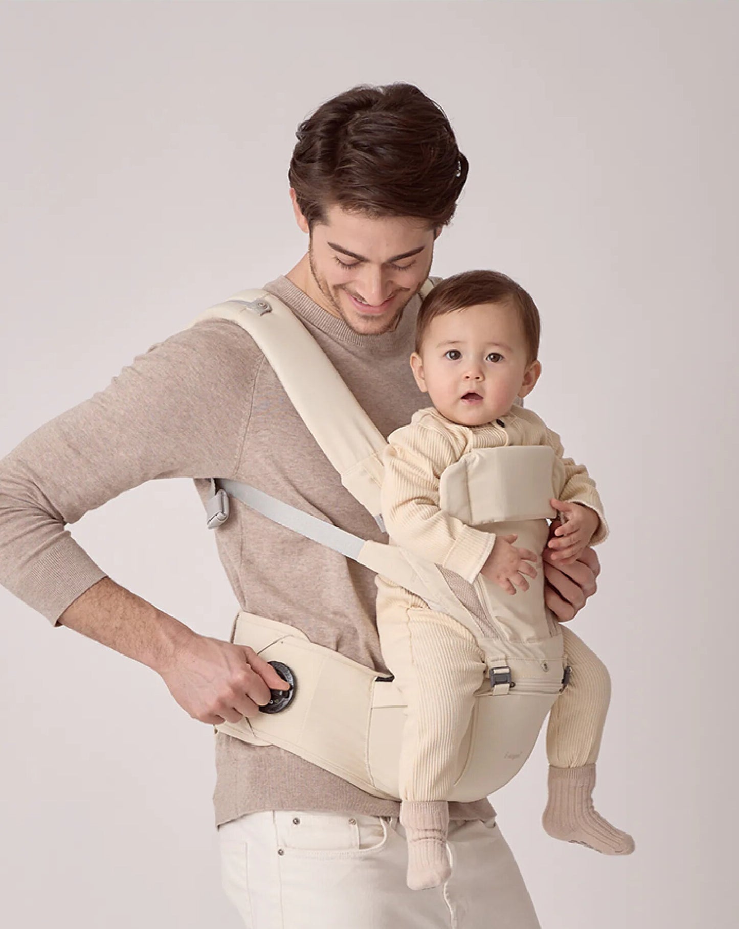 Man holding a baby in a beige baby carrier against a plain background showing the Dr Dial tightening system